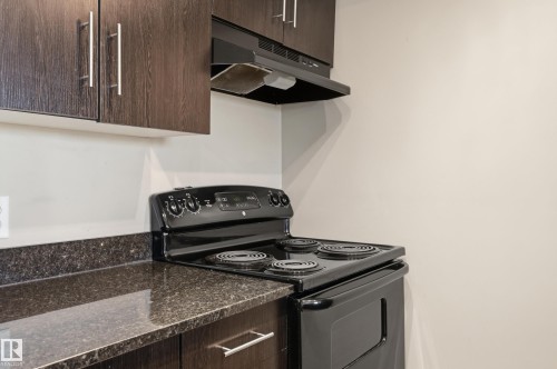 Kitchen featuring dark wood-finish cabinetry with silver hardware, speckled dark countertops, and a black electric range with an overhead vent - 304 11804 22 Avenue, Edmonton, AB 