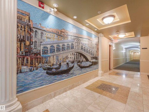 Corridor featuring an expansive wall mosaic art piece, decorative fluted column, coffered ceiling with recessed lighting and dome fixtures, and patterned tile flooring - 409 10333 112 Street, Edmonton, AB 