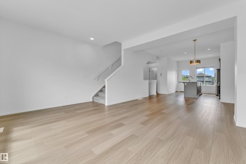 Open concept living area featuring light wood-finish flooring, a contemporary kitchen island with an undermount sink, a modern pendant light fixture, and a staircase with a white railing - 1534 157 Street, Edmonton, AB 
