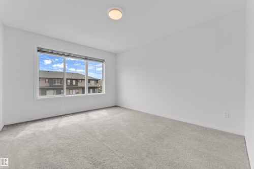 Spacious room featuring a large multi-pane window, neutral carpet flooring, white painted walls, white baseboards, and a flush-mount ceiling light fixture - 1534 157 Street, Edmonton, AB 