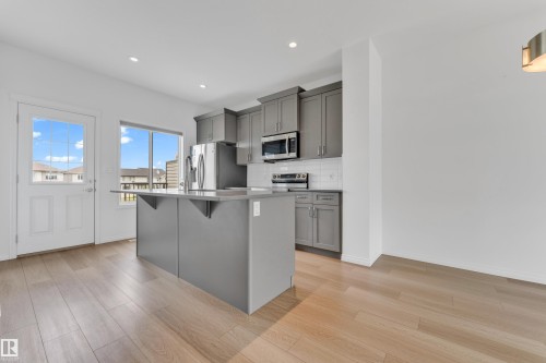 Modern kitchen featuring an island with a breakfast bar, stainless steel appliances, grey cabinetry, white subway tile backsplash, and wood-finish flooring - 1534 157 Street, Edmonton, AB 