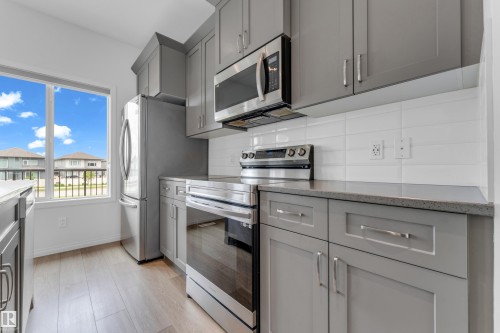 Modern kitchen featuring stainless steel appliances, grey cabinetry, light-toned wood-finish flooring, and a subway tile backsplash - 1534 157 Street, Edmonton, AB 