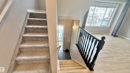 Split-level entry featuring light-toned wood-finish flooring, a black-painted banister, and a large window with grey drapes - 9 225 Blackburn Drive E, Edmonton, AB 