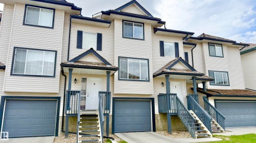 Multi-level townhome exterior featuring light-toned horizontal siding, dark trim, an attached garage with a panel door, a covered entry with a natural wood shingle accent roof, and a white front door - 9 225 Blackburn Drive E, Edmonton, AB 