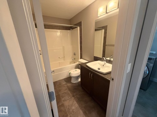 Bathroom featuring a white tub/shower combination, dark brown vanity with an integrated sink, and square tile flooring - 6821 Evans Wynd, Edmonton, AB 