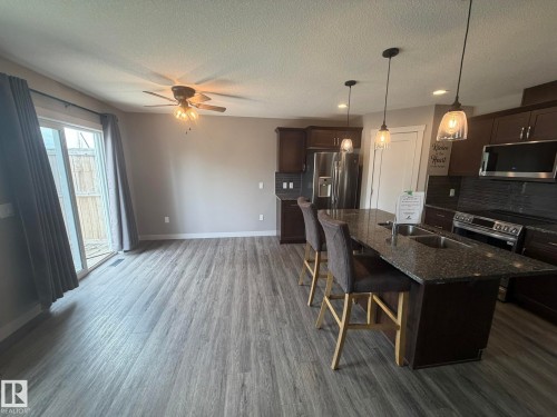 Open-concept kitchen and dining area featuring wood-finish flooring, a kitchen island with stone countertop and integrated double sink, dark wood cabinetry, stainless steel appliances, and sliding patio doors - 6821 Evans Wynd, Edmonton, AB 