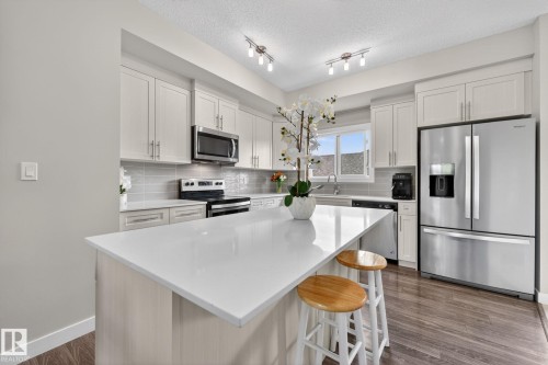 Bright kitchen featuring a white quartz countertop island, light-toned cabinetry, stainless steel appliances, a tiled backsplash, and wood-finish flooring - 165 Robinson Drive, Leduc, AB 
