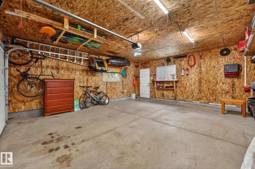 Attached garage featuring concrete flooring, exposed wood-finish walls and ceiling, overhead fluorescent lighting, a white pedestrian door, and built-in wall shelving - 165 Robinson Drive, Leduc, AB 