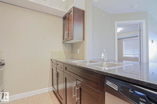Kitchen featuring dark wood cabinetry, granite-style countertops, double basin stainless steel sink with a chrome faucet, and integrated black dishwasher - 405 11804 22 Avenue Sw, Edmonton, AB 