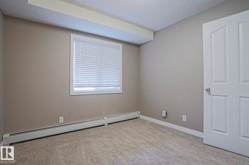Neutral-toned room featuring a window with blinds, baseboard heating, textured carpet flooring, and a paneled interior door - 405 11804 22 Avenue Sw, Edmonton, AB 