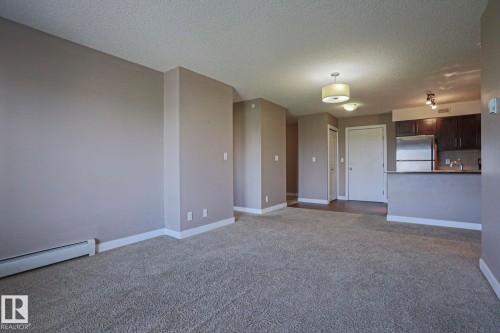 Open-concept living area featuring neutral-toned carpeting, light gray wall paint, and white baseboards - 405 11804 22 Avenue Sw, Edmonton, AB 
