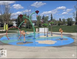 Community splash park featuring a circular concrete pad with colorful painted sections, multiple water play structures in varying designs, and surrounding green spaces - 