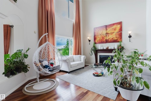 Living area featuring soaring ceilings, a fireplace with a dark wood mantel, wood-finish flooring, large windows, and wall sconces - 20524 58 Avenue, Edmonton, AB 
