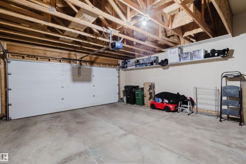 Attached garage featuring a white sectional garage door with an automatic opener, exposed wood ceiling joists, overhead lighting, a concrete floor, and an integrated storage shelf - 20524 58 Avenue, Edmonton, AB 