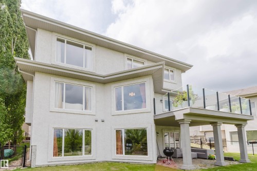 Contemporary stucco exterior with multiple large windows, a raised deck featuring glass railings, a covered patio area with structural columns, and a manicured lawn - 20524 58 Avenue, Edmonton, AB 