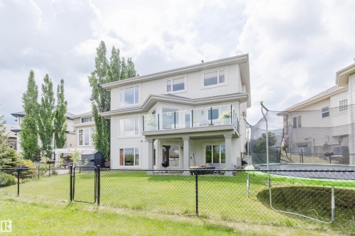 Expansive backyard featuring a manicured lawn, a two-story stucco exterior, a second-story balcony with a glass railing, a covered patio on the lower level, and mature perimeter trees - 20524 58 Avenue, Edmonton, AB 