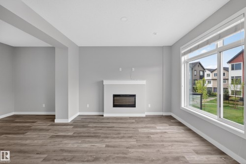 Spacious interior room with wood-finish flooring, a contemporary fireplace with a white mantel, and large windows providing abundant natural light - 90 1010 Rabbitt Hill Road Sw, Edmonton, AB - Indoor With Fireplace