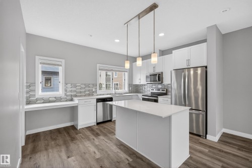 Modern kitchen featuring a central island with a light-toned countertop, stainless steel appliances, white cabinetry, wood-finish flooring, and a unique geometric tile backsplash - 90 1010 Rabbitt Hill Road Sw, Edmonton, AB - Indoor Photo Showing Kitchen With Stainless Steel Kitchen With Upgraded Kitchen