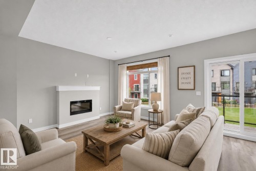 Living area featuring a contemporary fireplace with a white mantel, light gray walls, wood-finish flooring, and a sliding glass door leading to an outdoor space - 90 1010 Rabbitt Hill Road Sw, Edmonton, AB - Indoor Photo Showing Living Room With Fireplace