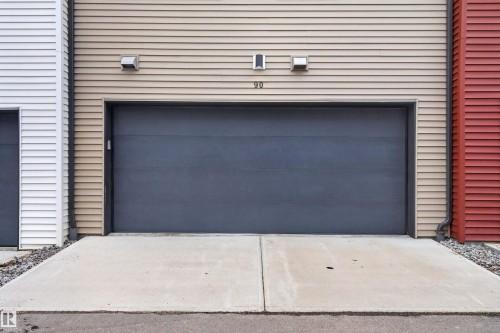 Expansive garage door with a concrete driveway, complemented by beige horizontal siding and exterior lighting fixtures - 90 1010 Rabbitt Hill Road Sw, Edmonton, AB - Outdoor With Exterior