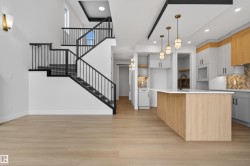 Open-concept layout featuring wood-finish flooring, a contemporary kitchen with light-toned cabinetry and a large island, a staircase with black metal railings, and integrated overhead lighting - 