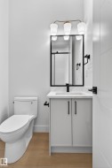 Contemporary bathroom featuring a white vanity with integrated sink, quartz-finish countertop, black framed mirror, three-light vanity fixture with opaque white shades, and wood-finish flooring - 