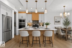 Kitchen and dining area featuring a central island with countertop seating, stainless steel appliances, light wood-finish flooring, light gray and wood-tone cabinetry, and a herringbone-patterned tile backsplash - 