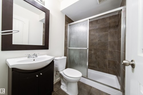 Bathroom featuring a dark wood-finish vanity with an integrated basin sink, a framed mirror, a toilet, a shower with sliding glass doors, and brown tile flooring - 405 Simmonds Way, Leduc, AB 