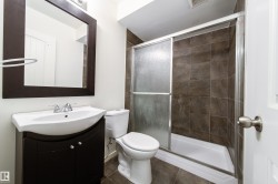 Bathroom featuring a dark wood-finish vanity with an integrated basin sink, a framed mirror, a toilet, a shower with sliding glass doors, and brown tile flooring - 
