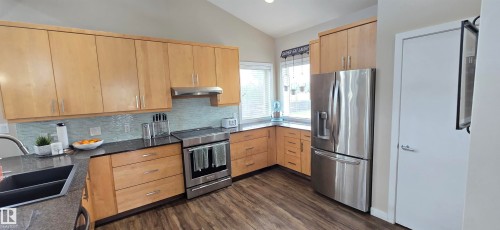 Contemporary kitchen featuring wood-finish cabinetry, stainless steel appliances, dark countertops, a light blue tiled backsplash, and wood-finish flooring - 4234 46 Street, Vegreville, AB - Indoor Photo Showing Kitchen With Stainless Steel Kitchen With Double Sink