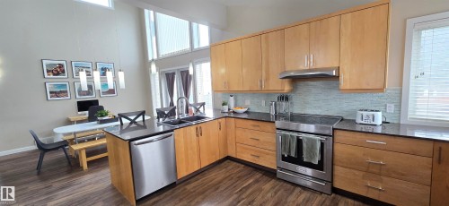Open-concept kitchen featuring wood-finish flooring, light wood cabinetry, dark countertops, and stainless steel appliances - 4234 46 Street, Vegreville, AB - Indoor Photo Showing Kitchen With Stainless Steel Kitchen With Double Sink