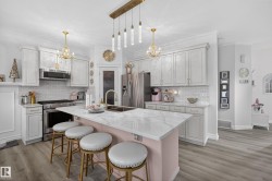 Spacious kitchen featuring an extended island with a white veined countertop, light gray cabinetry with gold-tone hardware, a tiled backsplash, stainless steel appliances, and wood-finish flooring - 