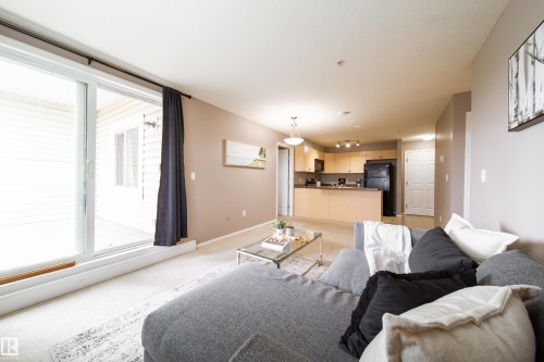 Open-concept living area featuring light-colored carpet, a sliding glass door providing access to an outdoor space, and a kitchen with wood-finish cabinetry and black appliances - 115 4309 33 Street, Stony Plain, AB 