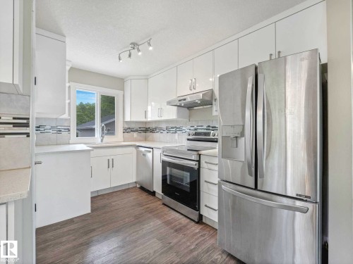 Modern kitchen featuring white cabinetry, stainless steel appliances, light-toned countertops, a mosaic tile backsplash, and wood-finish flooring - 12241 54 Street, Edmonton, AB 