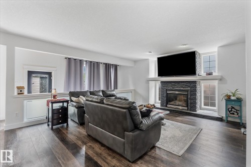 Living area featuring wood-finish flooring, a stone-clad fireplace with a light-colored mantel, a built-in television mount, recessed lighting, and a window with gray drapes - 18 Woodbend Way, Fort Saskatchewan, AB 