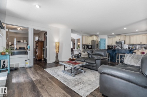Open-concept living area featuring wood-finish flooring, recessed lighting, and a kitchen with cream-colored cabinetry, a dark tile backsplash, and a blue-paneled island with seating - 18 Woodbend Way, Fort Saskatchewan, AB 
