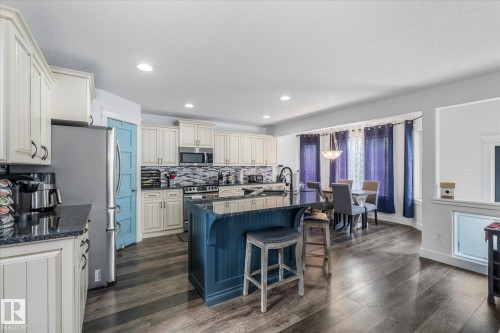 Kitchen featuring a central island with seating, dark countertops, white cabinetry, stainless steel appliances, and wood-finish flooring - 18 Woodbend Way, Fort Saskatchewan, AB 