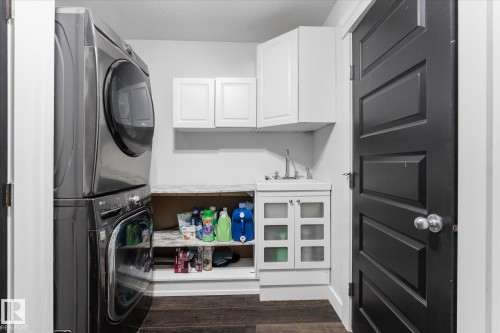 Dedicated laundry area featuring stacked appliances, a utility sink with a vanity, upper and lower cabinetry, wood-finish flooring, and a paneled interior door - 18 Woodbend Way, Fort Saskatchewan, AB 