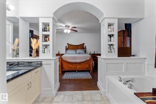 Arched entryway connecting spaces, featuring built-in shelving, a deep soaking tub with chrome fixtures, a vanity with a dark speckled countertop, and light-toned cabinetry - 18 Woodbend Way, Fort Saskatchewan, AB 