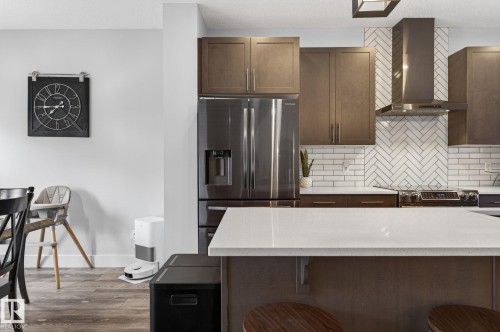 Modern kitchen featuring an expansive island with a light-toned countertop, a stainless steel range hood, dark wood-finish cabinetry, a herringbone pattern tile backsplash, and wood-finish flooring - 17733 64A Street, Edmonton, AB 