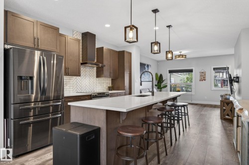 Modern kitchen featuring wood-finish cabinetry, stainless steel appliances, a white countertop island with seating, pendant lighting, and wood-finish flooring - 17733 64A Street, Edmonton, AB 