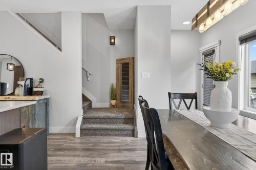 Open-concept dining area featuring wood-finish flooring, a contemporary linear light fixture, a staircase with carpeted treads and white risers, a wood-finish dining table, and a wall-mounted sconce - 17733 64A Street, Edmonton, AB 