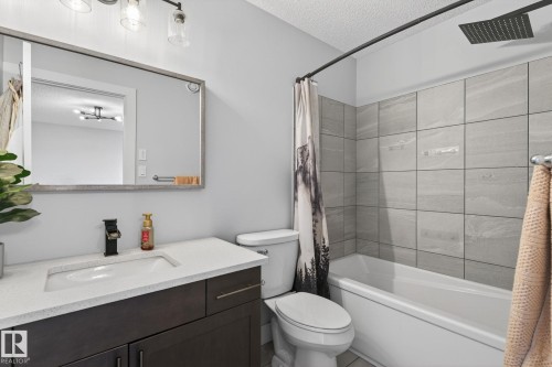 Full bathroom featuring a dark wood-finish vanity with a white countertop and integrated sink, a rectangular framed mirror, and a full-size bathtub with gray tile surround and a matte black rainfall showerhead - 17733 64A Street, Edmonton, AB 
