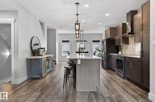 Open-concept kitchen featuring a central island with countertop seating, wood-finish flooring, dark wood cabinetry, stainless steel appliances, and a tiled backsplash - 17733 64A Street, Edmonton, AB 
