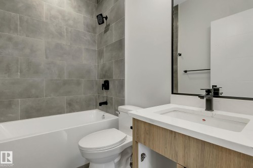 Bathroom featuring a tub/shower combination with large format grey tile surround, matte black fixtures, a wood-finish vanity with an integrated sink, and a framed mirror - 11132 72 Avenue, Edmonton, AB - Indoor Photo Showing Bathroom