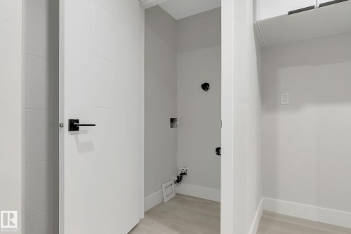 Utility area featuring light-toned flooring, white baseboards, a white paneled door with matte black hardware, and an overhead white cabinet - 11132 72 Avenue, Edmonton, AB - Indoor