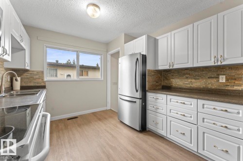 Kitchen featuring white shaker-style cabinetry, wood-finish flooring, a stainless steel refrigerator, a double basin sink with a gooseneck faucet, and a tiled backsplash - 210 22 Street, Cold Lake, AB 
