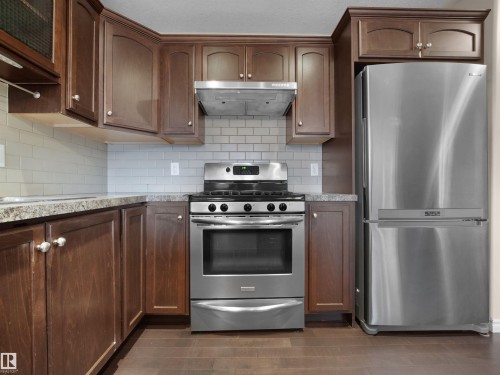 Kitchen featuring wood-finish flooring, dark wood cabinetry, a subway tile backsplash, stainless steel appliances, and stone-look countertops - 21316 45A Avenue Nw, Edmonton, AB 