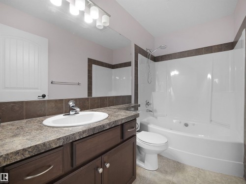 Bathroom featuring a white oval sink set into a speckled countertop, dark wood-finish cabinetry, a white bathtub with a built-in shower, brown tile accents, and a white toilet - 21316 45A Avenue Nw, Edmonton, AB 