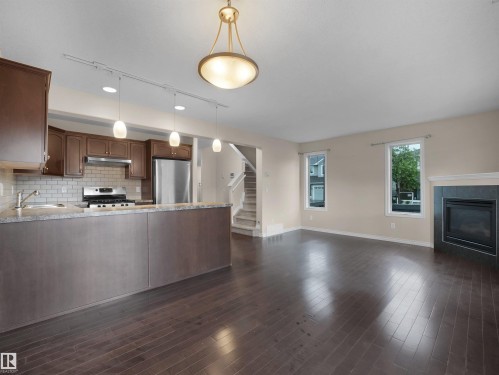Open-concept living space featuring wood-finish flooring, a kitchen island with stone-finish countertops, stainless steel appliances, a subway tile backsplash, and a fireplace with a mantel - 21316 45A Avenue Nw, Edmonton, AB 